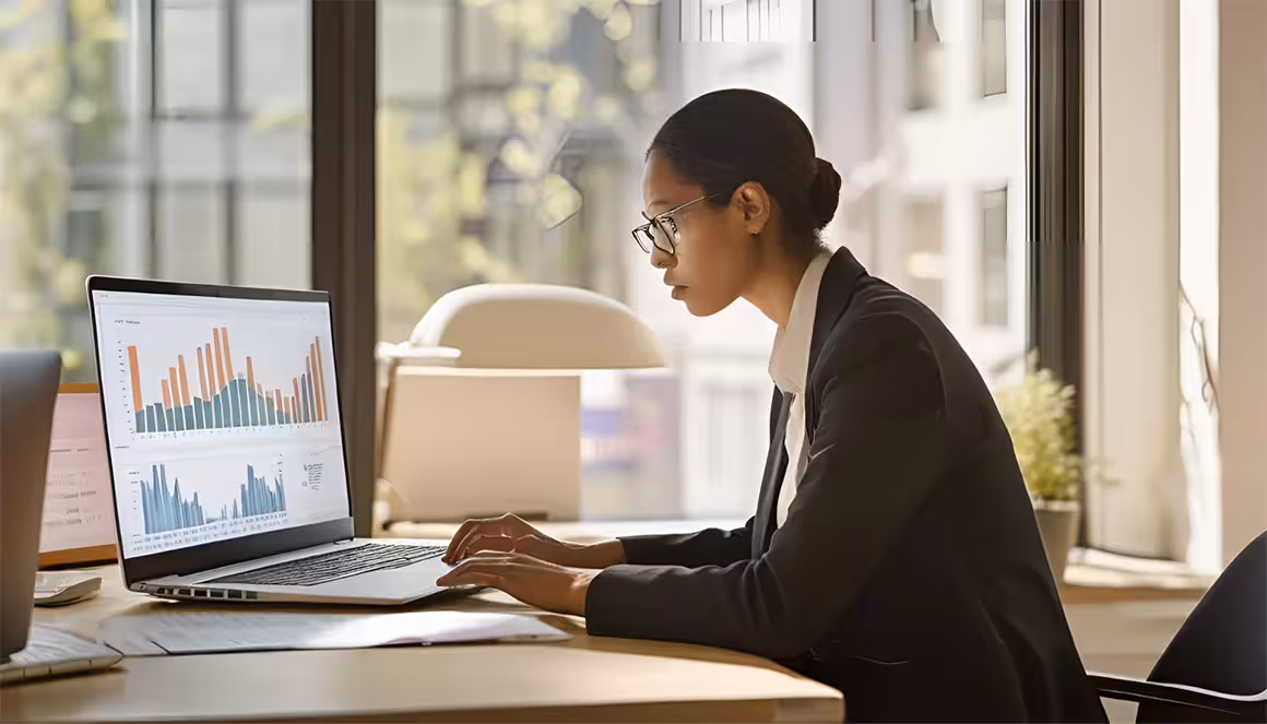 A focused professional woman in a suit works at a laptop displaying colorful bar charts using flow data tools. Sunlit office with large windows. Analytical and productive mood.